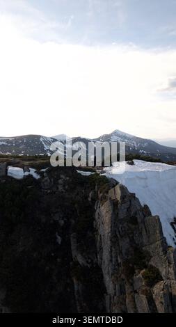 Drohnenperspektive von zwei winzigen Figuren, die auf einem dramatischen felsigen Kamm mit dicken Schneegesimsen vor dem Hintergrund majestätischer Berggipfel stehen Stockfoto