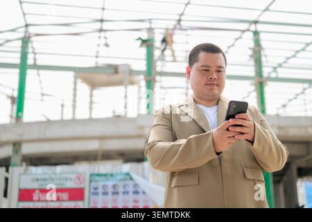 Eine kleine Aufnahme eines fokussierten mittelgroßen asiatischen Geschäftsmannes aus der Region Isan, der auf seinem Handy vor einem Gebäudegerüst unter c schreibt Stockfoto