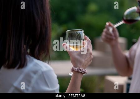 Eine junge Frau, die von hinten gesehen wird, hält ein stielloses Glas Weißwein, während ein Begleiter ein Glas Rotwein in einem leicht verschwommenen Garten hält Stockfoto