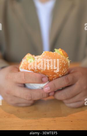 Eine Nahaufnahme konzentriert sich auf die Hände eines großen mittelgroßen asiatischen Mannes, der einen gebissenen Sesamhamburger über einem Holztisch mit einer flachen Ablage hält Stockfoto