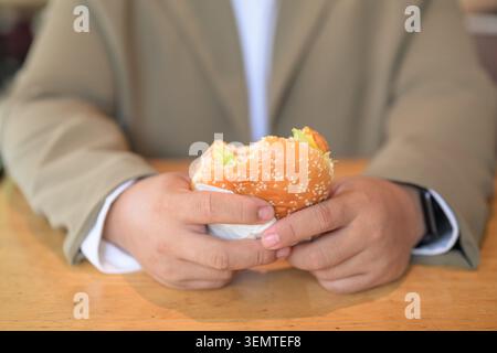 Eine Nahaufnahme konzentriert sich auf die Hände eines großen mittelgroßen asiatischen Mannes, der einen gebissenen Sesamhamburger über einem Holztisch mit einer flachen Ablage hält Stockfoto