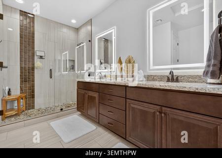 Helles, modernes Badezimmer mit dunklem Holz-Waschtisch mit Granitablagefläche, gerahmten Spiegeln mit Hintergrundbeleuchtung, rahmenloser Glasdusche und beigefarbenen Tönen Stockfoto