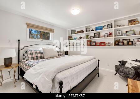 Helles Schlafzimmer mit schwarzem Metallrahmen, weißer Bettwäsche mit karierten Kissen, einer Reihe eingebauter weißer Regale mit gerahmten Fotos und Deko Stockfoto