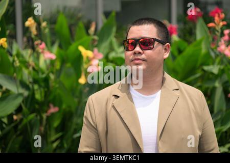 Ein nachdenklicher junger Erwachsener aus der Isan-Region steht draußen und blickt nachdenklich weg, während er eine rote reflektierende Sonnenbrille trägt Stockfoto