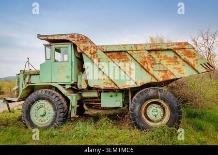14. April 2026, Izvor, Serbien: Ein schwerer Muldenkipper mit Rost und Verfall, der in Gras und Unkraut unter blauem Himmel sitzt und die Aufgabe A repräsentiert Stockfoto