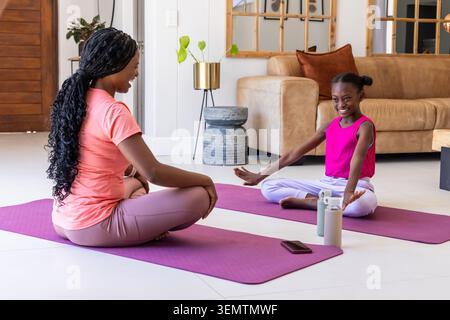 Afroamerikanische Mutter und Jugendtochter machen Yoga auf lila Matten im Wohnzimmer mit Smartphone Stockfoto