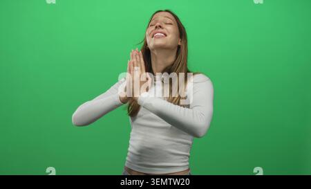 Junge hispanische Frau mit zusammengedrückten Händen betend im grünen Atelier, Augen geschlossen und ein leichtes Lächeln; Hoffnung. Stockfoto