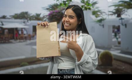 Die junge Frau lächelt und zeigt mit dem Finger auf eine braune Papiertüte, während sie sie in einer belebten Fußgängerzone in der Brust hält; fröhliche Beförderung. Stockfoto