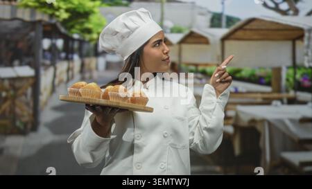 Die junge Küchenchefin hält ein Tablett mit Muffins und zeigt mit dem Finger an den Verkaufsständen auf dem Straßenmarkt; Selbstvertrauen stolz auf Handwerkskunst Wärme. Stockfoto