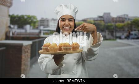 Die junge hispanische Chefin lächelt und hält ein hölzernes Tablett mit Muffins in der Hand, während sie mit dem Finger auf Muffins auf einer Straße der Stadt zeigt; fröhlicher Stolz. Stockfoto