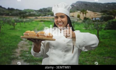 Die junge Küchenchefin hält ein hölzernes Tablett mit Muffins und gibt die Daumen vor dem Gebäude hoch; stolz auf das Backen. Stockfoto