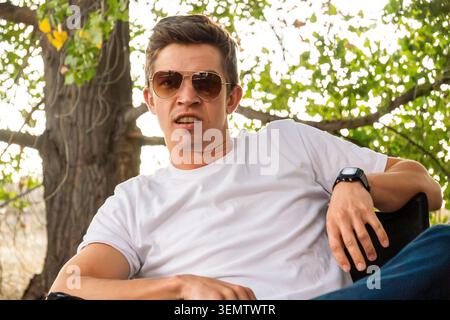 Ein Mann sitzt draußen in einem Park unter grünen Bäumen, trägt ein weißes kurzärmeliges T-Shirt und eine Sonnenbrille, mit einer entspannten, selbstbewussten Pose an natürlichem Tag Stockfoto