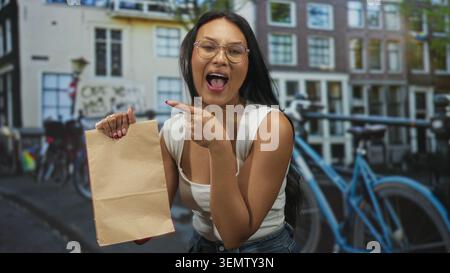 Die junge Frau hält eine braune Papiertüte und zeigt mit dem Finger, während sie mit dem Fahrrad auf der Stadtstraße lächelt; lässige Beförderungsfreude. Stockfoto