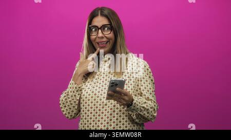 Frau, die das Smartphone hält und den Finger nach oben zeigt, während sie im Studio eine große Brille trägt; verspielte Neugier. Stockfoto
