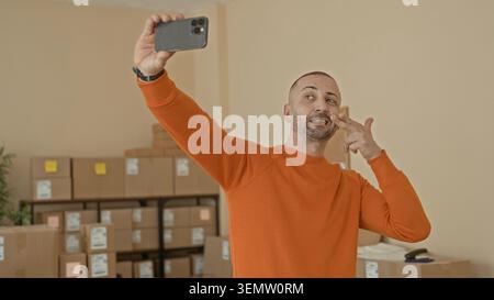 Ein Mann im orangefarbenen Pullover hält das Smartphone für Selfie und zeigt mit der Hand ein Friedenszeichen zwischen gestapelten Kartonschachteln in einem Gebäude; Stolz auf Kleinunternehmen. Stockfoto