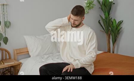 Mann, der am Rande eines Bettes sitzt und gähnt, während er sich in einem Studio den Hals dehnt und reibt; Morgenroutine schläfrig. Stockfoto