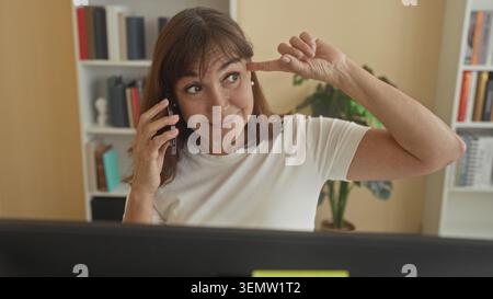 Frau, die auf dem Smartphone spricht, zeigt mit dem Finger auf den Tempel im Büro, wobei der Computerbildschirm sichtbar ist; nachdenklicher Fokus. Stockfoto