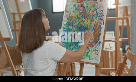 Malerin steht an der Staffelei, rechte Hand malt dicke bunte Striche auf Leinwand, während sie Palette hält, Rückansicht im Kunststudio; Konzentration cr Stockfoto