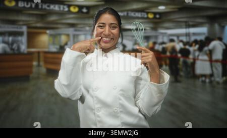Die Küchenchefin, die den Schneebesen hält, zeigt in der Tickethalle des Flughafens mit dem Finger auf die Zähne, lächelt und zeigt Zähne; Stolz-Feier. Stockfoto