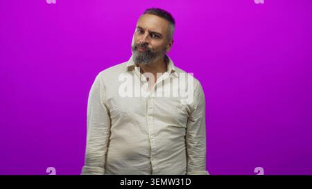 Mann mit grauem Bart, Sträucher und Spitzen mit offenen Handflächen in lila Studio, mit beigem Hemd und perlenbesetzten Armbändern; Ungewissheit. Stockfoto