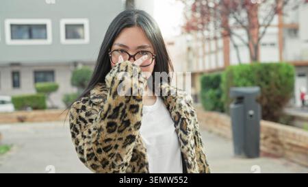 Frau zeigt den Finger nach vorne, trägt Mantel und Brille auf der Straße mit Leopardenmuster, lächelt und engagiert sich mit der Kamera; freundliches Vertrauen. Stockfoto