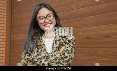 Frau mit Leopardenmantel und Brille lächelt mit überkreuzten Armen vor dem Gebäude; Vertrauenswärme. Stockfoto