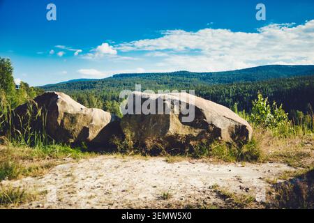 Zwei große Felsbrocken auf einem sonnendurchfluteten Boden in den Sudetes Mountains, Polen, die ein bewaldetes Tal und sanfte Hügel unter einem hellblauen Himmel überblicken Stockfoto