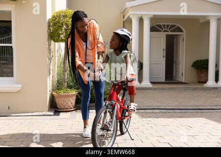 Afroamerikanische Mutter und Kind üben auf rotem Fahrrad in der Einfahrt, während Kind Helm trägt Stockfoto
