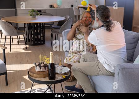 Ein älteres Paar sitzt auf einem grauen Sofa, während ein Mann einen gelben Partyhut neben Kuchen und Sektflöten legt Stockfoto