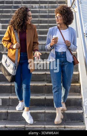 Afroamerikanische Frauen in Blazern, die mit der Schiene Betonstufen hinunterlaufen, mit einer Tasche, in der Kaffee aufbewahrt wird Stockfoto