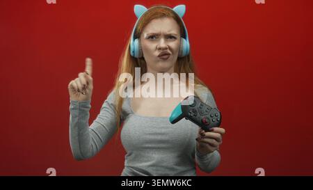 Junge rothaarige Frau, die mit dem Finger zeigt, während sie ein blaues Gamepad hält und Katzenohrkopfhörer in einem Studio mit rotem Hintergrund trägt; Frustrationsherausforderung. Stockfoto