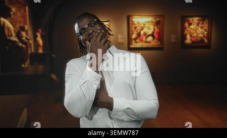Mann mit Hand über Mund und Daumen auf der Brust in der Galerie mit weißem Hemd und Brille unter warmer Beleuchtung; amüsante Reflexion. Stockfoto
