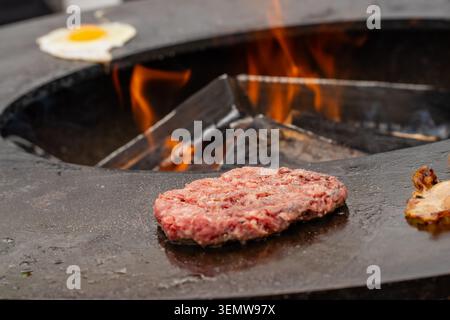 Frisches Fleischschnitzel und ein ganzes Ei-Braten auf einer runden Grillstation aus Metall, wobei brennende Holzscheite Rauch während des Cateringservice im Freien ausstoßen Stockfoto