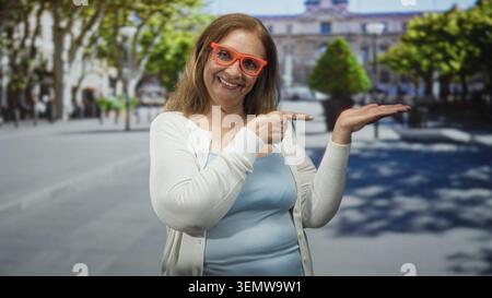 Frau, die mit dem Finger auf eine belebte Straße zeigt, trägt eine orange Brille und ein hellblaues Tanktop; fröhliche Einladung. Stockfoto