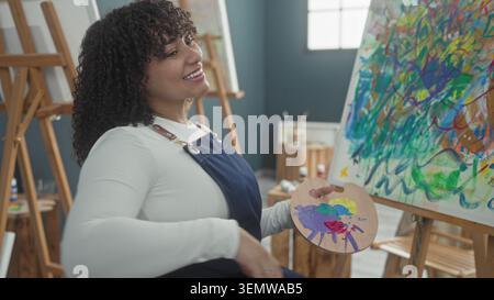 Die Malerin hispanic Brünette hält eine hölzerne Farbpalette und einen Pinsel, während sie lächelt und in einem Kunststudio, cre, bunte Striche auf eine Leinwand aufträgt Stockfoto