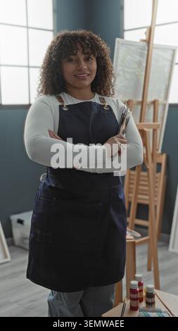 Malerin, die Pinsel mit gekreuzten Armen in einem Atelier hält, in marineblauer Schürze und weißem Hemd; ruhiges Vertrauen. Stockfoto