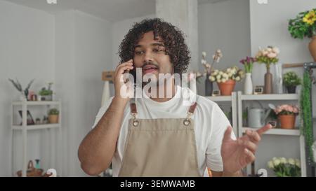 Mann, der mit Handgesten auf dem Smartphone spricht, lächelnd, Florist trägt Schürze zwischen Blumenregalen in einem Gebäude; Service ruhig. Stockfoto