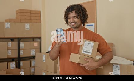 Mann in orangefarbenem Hemd hält Kartons und Smartphone, lächelt, während er in einem Pappraum des Gebäudes telefoniert; Optimismus für kleine Unternehmen. Stockfoto