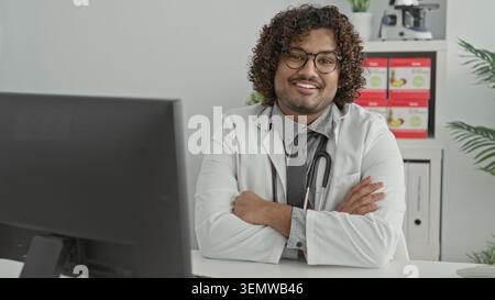 Mann Doktor mit Stethoskop, Arme überkreuzt am Klinikpult, lächelt in die Kamera, Mitgefühl Selbstvertrauen. Stockfoto