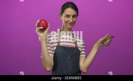 Frau, die roten Apfel in Handschuhen hält und in lila Studio zur Seite zeigt, mit gestreiftem Hemd und Denim Schürze; gesunde Ernährung fröhlich. Stockfoto