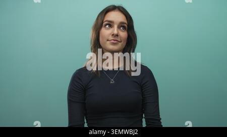 Junge hispanische Frau mit Herzkette blickt im Studio vor blauem Hintergrund auf; nachdenkliche Reflexion. Stockfoto