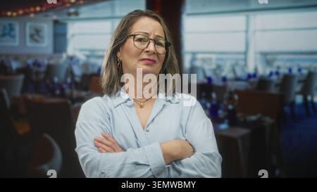 Hispanische Seniorin mit grauen Haaren, Brille und Halskette, überkreuzten Armen und einer Stirnrunze in einem Restaurantgebäude; Ärgernis. Stockfoto