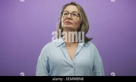 Frau hispanic Senior mit grauem Haar und Brille zeigt den Zeigefinger nach oben, während sie vor einem violetten Studio-Hintergrund aufblickt; Neugier. Stockfoto
