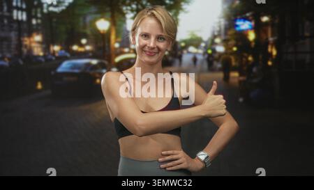 Frau lächelt mit Händen auf den Hüften und exponierten bauchmuskeln auf der Stadtstraße bei Dämmerung unter der Abendlaterne; Selbstvertrauen Wohlbefinden. Stockfoto