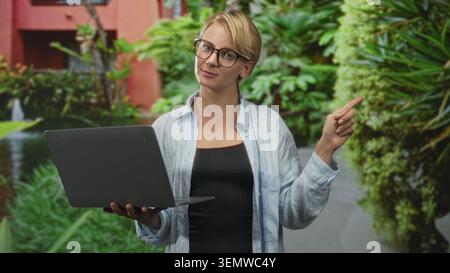 Blonde Frau mit Brille, die den Laptop hält, zeigt mit dem rechten Zeigefinger in den Innenhof mit grünen Pflanzen und Brunnen; gute Orientierungshilfe Stockfoto