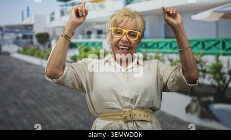 Eine ältere hispanische Frau mit gelber Brille und Armbändern lächelt und zeigt mit den Zeigefingern nach oben, während sie ein gürteltes Leinenhemd vor einem Gebäude trägt Stockfoto