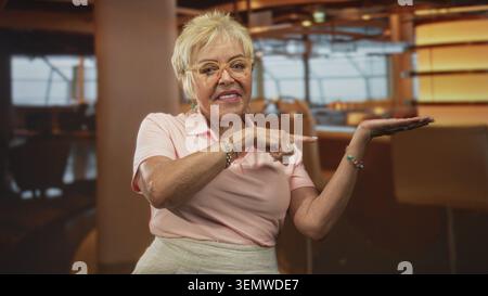 Die ranghöchste hispanische Frau zeigt mit dem Finger auf die Handfläche in der Flughafenlounge, trägt Brillen und Armbänder; fröhliches Vertrauen. Stockfoto