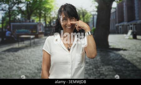 Eine ältere Frau zeigt beim Lächeln mit dem Finger an die Nase, trägt weiße Bluse und Anhänger auf einer Kopfsteinpflasterstraße. Stockfoto