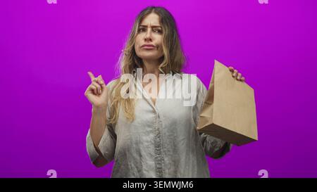Frau zeigt mit dem Finger und hält die braune Papiertüte vor violettem Studio-Hintergrund, während sie nach links drückt; Frustration. Stockfoto