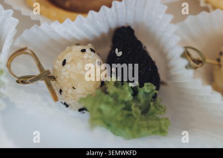 Zarte Reisbällchen ruhen in einem knusprigen weißen Becher. Schwarze und weiße Sesamkörner sind auf jeden Ball gestopft. Frischer grüner Salat sorgt für einen lebendigen Kontrast. Goldener Draht t Stockfoto